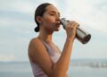 A woman drinks from a metal bottle on a sunny day by the beach, wearing a sports bra.
