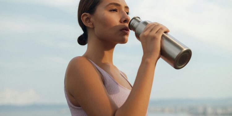 A woman drinks from a metal bottle on a sunny day by the beach, wearing a sports bra.