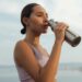 A woman drinks from a metal bottle on a sunny day by the beach, wearing a sports bra.